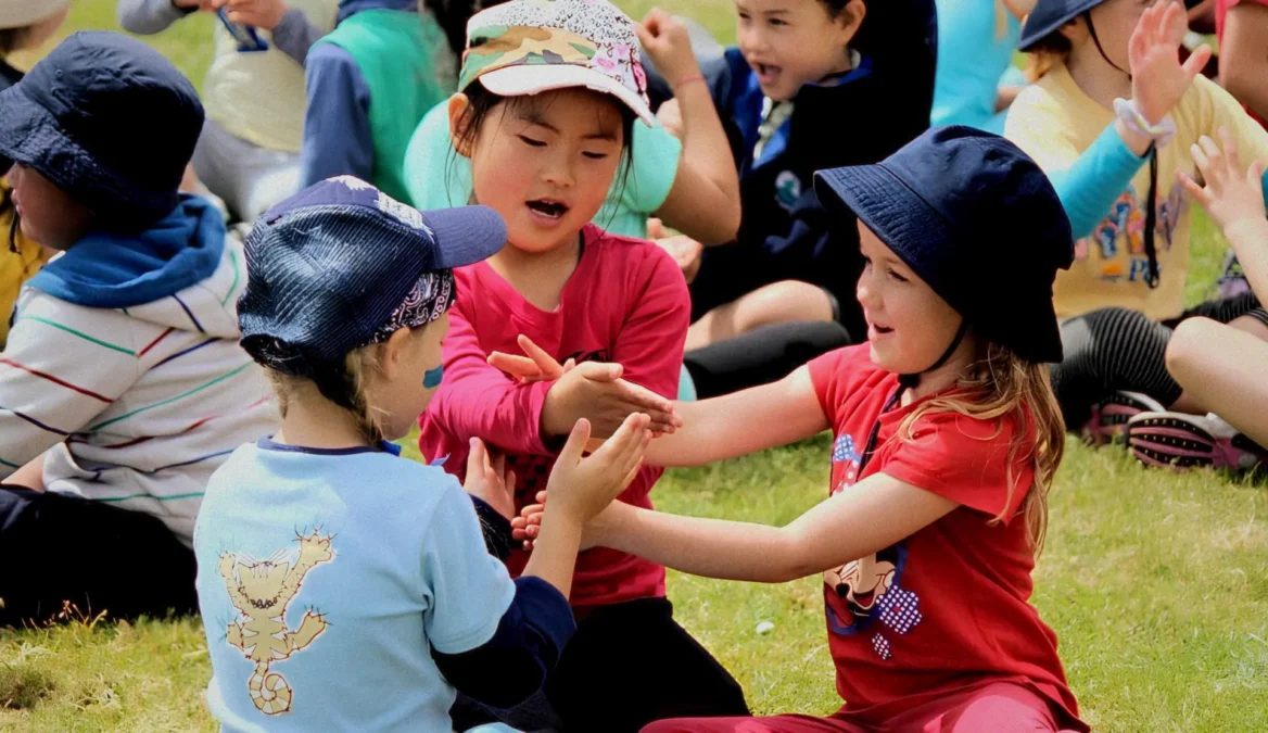 A photo of three girls with sunhats playing patty-cake on a field.