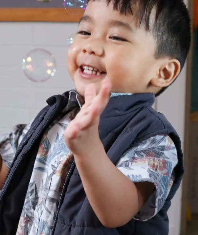 A toddler giggles excitedly, reaching out to touch bubbles floating around him during a children's activity at church.