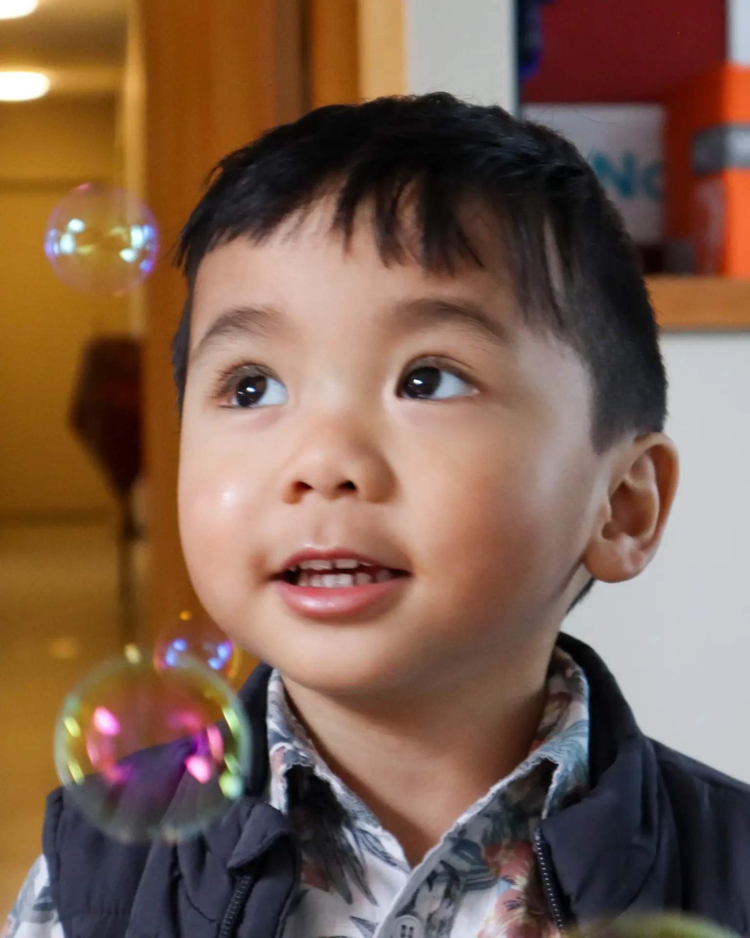 A filipino toddler with short hair in a vest jacket, looking curiously at bubbles as they fall down slowly.