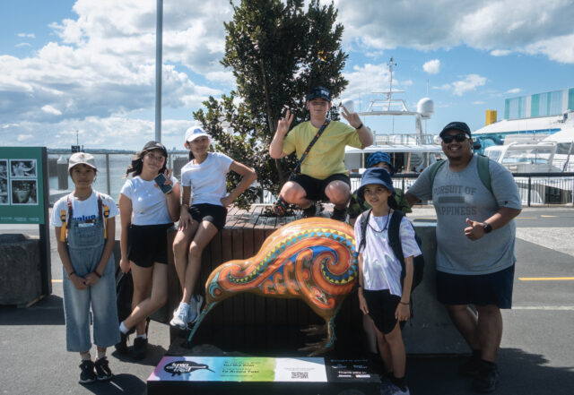 A group of pre-teens with an adult, smiling at the camera while seated on a tall planter box. In front of them is a kiwi statue with bright colours and traditional Maori design.