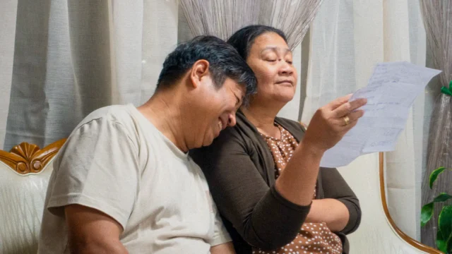 A close-up of an elderly Asian couple sitting on a couch in a living room. The woman, wearing a brown and polka-dot top, holds a piece of paper and reads with her eyes closed in concentration, while the man leans his head against her shoulder and smiles warmly.
