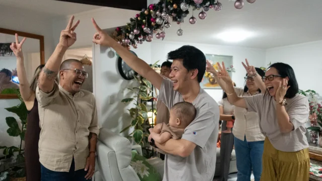 A joyful group of diverse adults celebrate indoors, with many raising their hands in victory signs and laughter. One man holds a baby while pointing upwards, and festive garlands are strung overhead.
