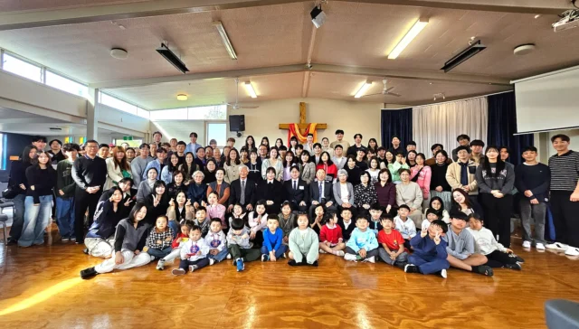 A large group photo of the Korean Evangelical Church congregation, featuring a diverse mix of adults and children smiling and posing together inside Glenfield Baptist Church, with a wooden cross.
