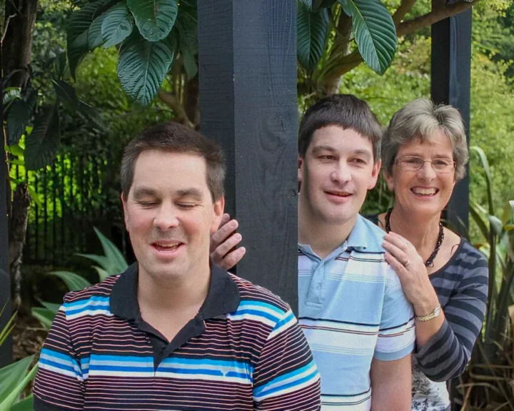 Three people, two men and an older woman, pose outdoors amongst lush green foliage. One man in a striped shirt has his eyes closed and is smiling, while another man peeks from behind a dark wooden post, with the smiling woman's hand on his shoulder.