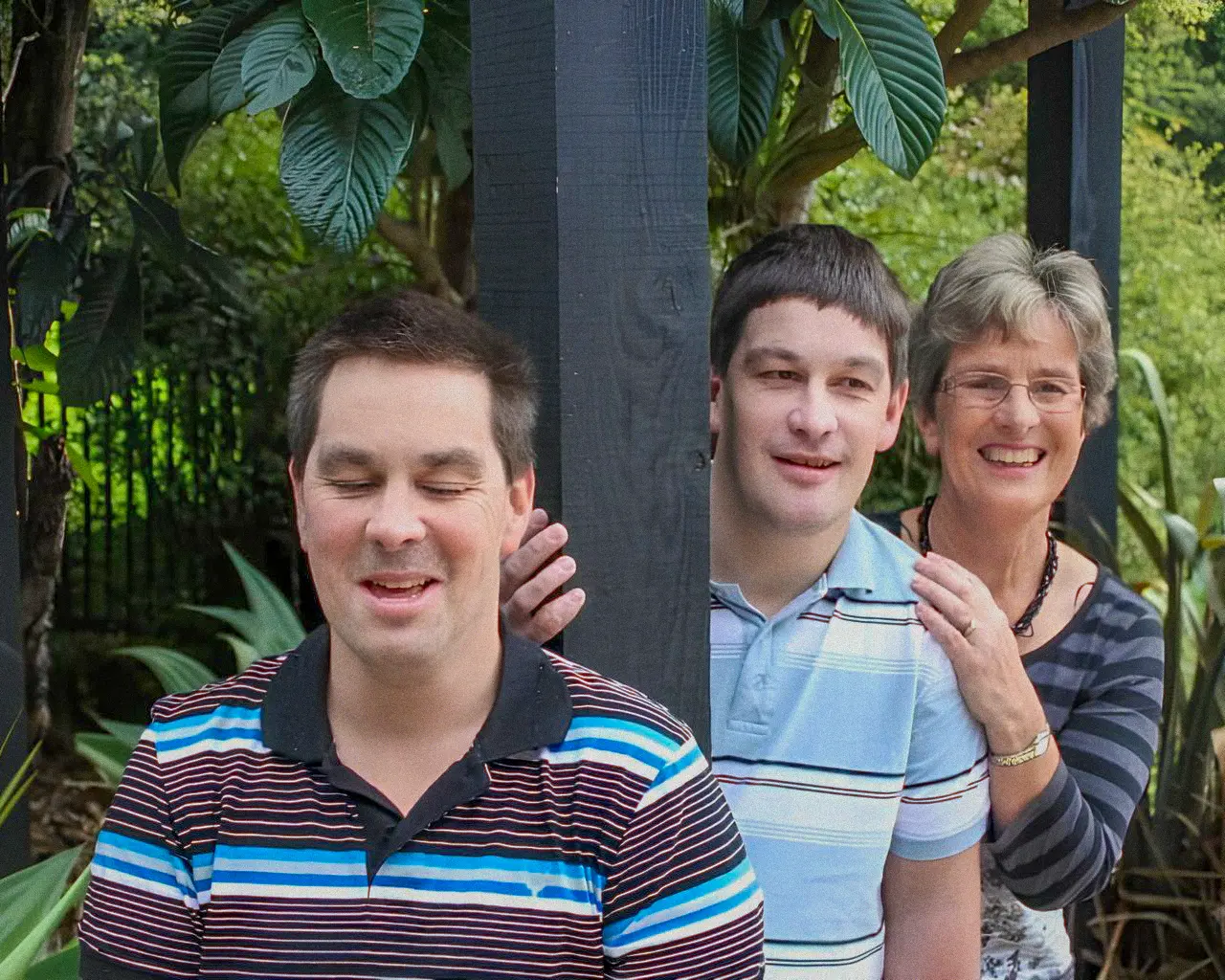 Three people, two men and an older woman, pose outdoors amongst lush green foliage. One man in a striped shirt has his eyes closed and is smiling, while another man peeks from behind a dark wooden post, with the smiling woman's hand on his shoulder.