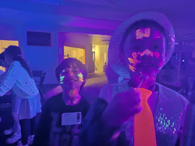 Two Asian children smiling at the camera in a dimly lit room. They have glow-in-the-dark face paint and attire.
