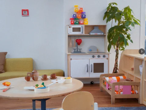 One of the play rooms at Little Nest Early Centre in Glenfield, North Shore. It features A cozy play area for children featuring a round wooden table with toy teacups and play food, a miniature wooden play kitchen set with colorful toy food stacked above it, and a small wooden doll crib, all in a room with a lime green sofa and a tall green plant.