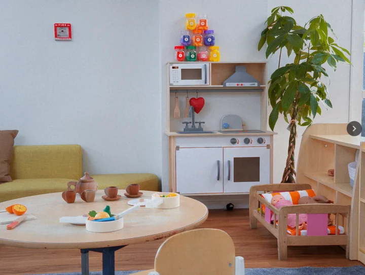 One of the play rooms at Little Nest Early Centre in Glenfield, North Shore. It features A cozy play area for children featuring a round wooden table with toy teacups and play food, a miniature wooden play kitchen set with colorful toy food stacked above it, and a small wooden doll crib, all in a room with a lime green sofa and a tall green plant.