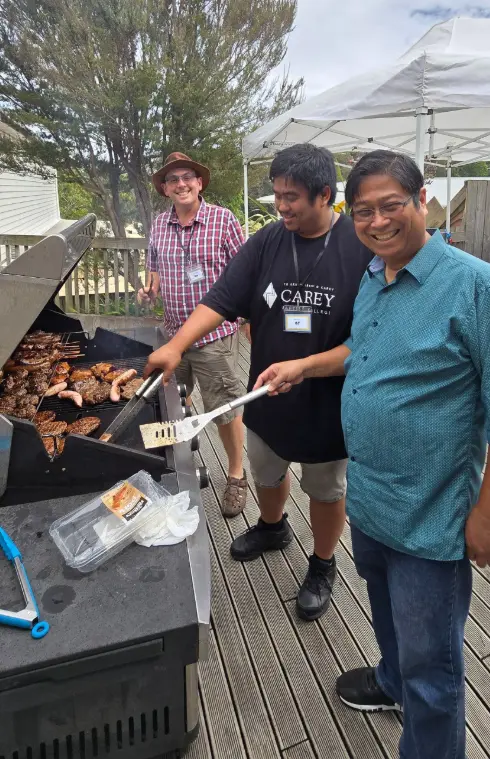 Two Filipino men and one Caucasian man smile at the camera, while cooking BBQ during a church event.