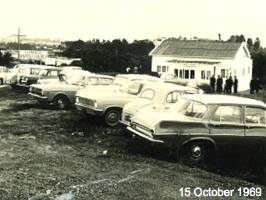 Black and white photograph, dated 15 October 1969, showing a line of vintage cars parked in a grassy field next to a small, single-story white building, with a few people standing by the entrance.