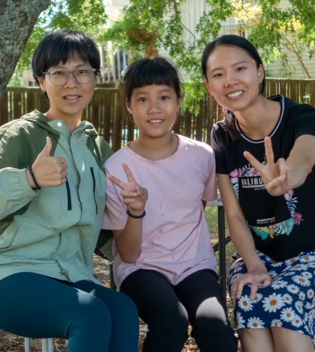 Three East Asian church members, two women and a girl, smile and make peace signs while seated outdoors at a church picnic.