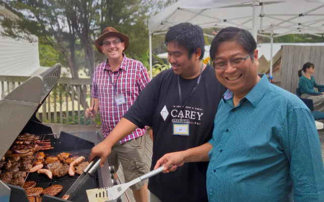 Two Filipino men and one Caucasian man smile at the camera, while cooking BBQ during a church event.