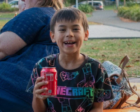 A boy smiling into the camera with a Coca-cola can in his hands as he sits on a picnic in the park.