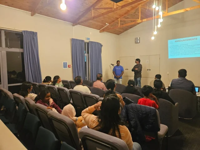 A photo of a North Shore Tamil Fellowship gathering at Glenfield Baptist Church. Two men stand at the front near a projection screen with "ANNOUNCEMENTS" visible, addressing a group of people seated in rows of auditorium-style chairs. The room has a wooden ceiling and is lit by overhead lights.