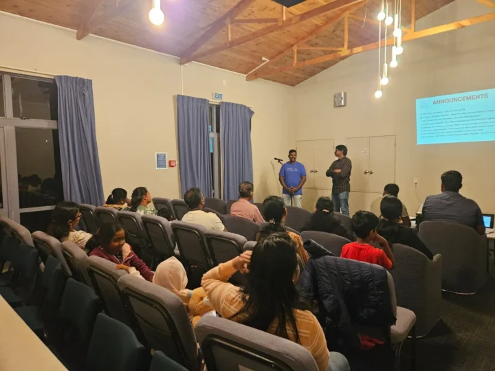 A photo of a North Shore Tamil Fellowship gathering at Glenfield Baptist Church. Two men stand at the front near a projection screen with "ANNOUNCEMENTS" visible, addressing a group of people seated in rows of auditorium-style chairs. The room has a wooden ceiling and is lit by overhead lights.