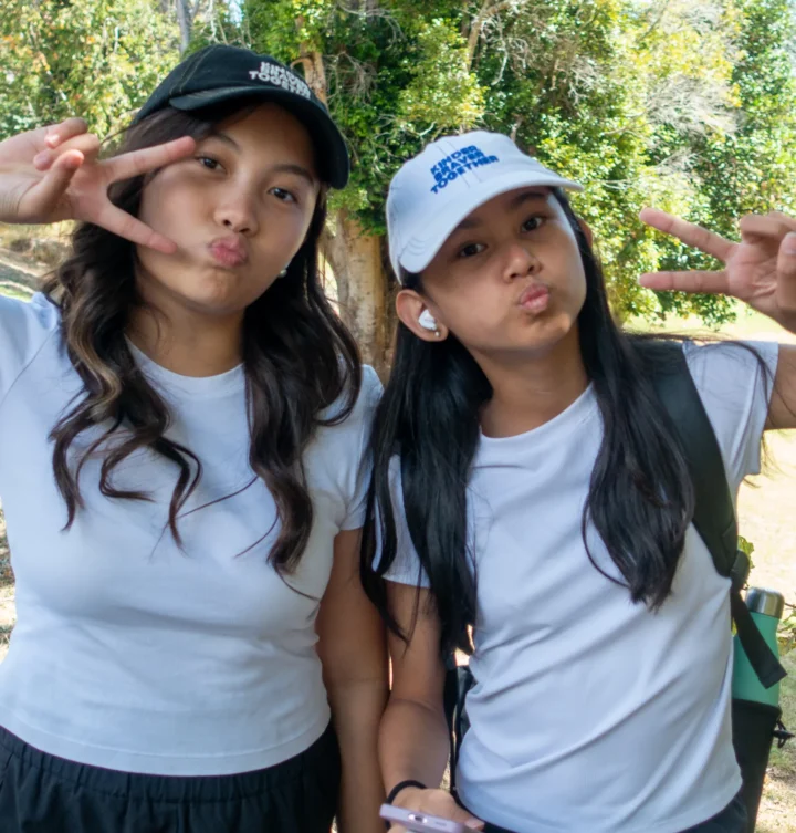 Query successful Two Asian girls from the church youth group wear matching white t-shirts and caps, pucker their lips, and make peace signs outdoors.