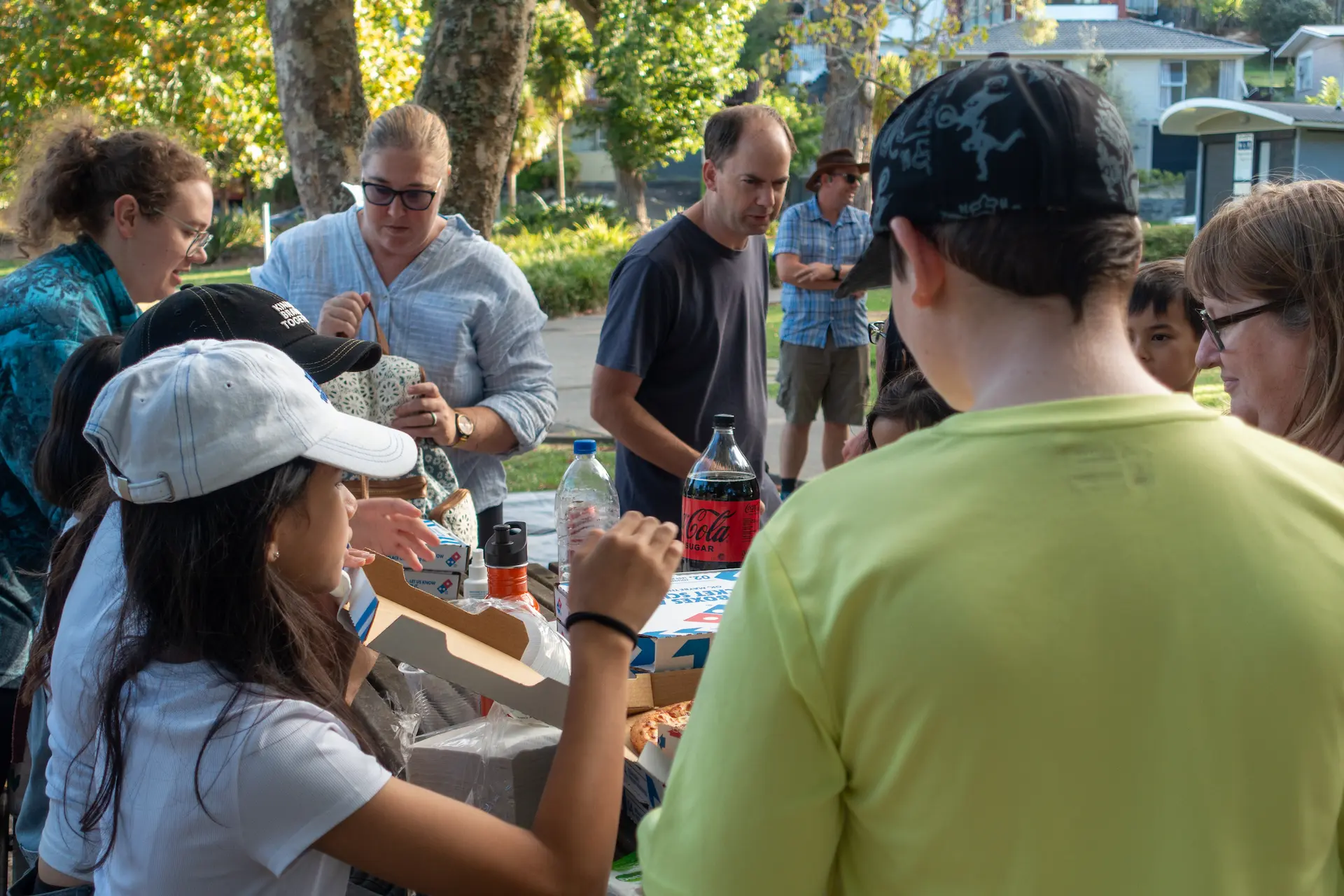 A group of diverse adults and children gathered around a picnic table in a park, reaching for food and drinks including pizza and Coca-Cola.